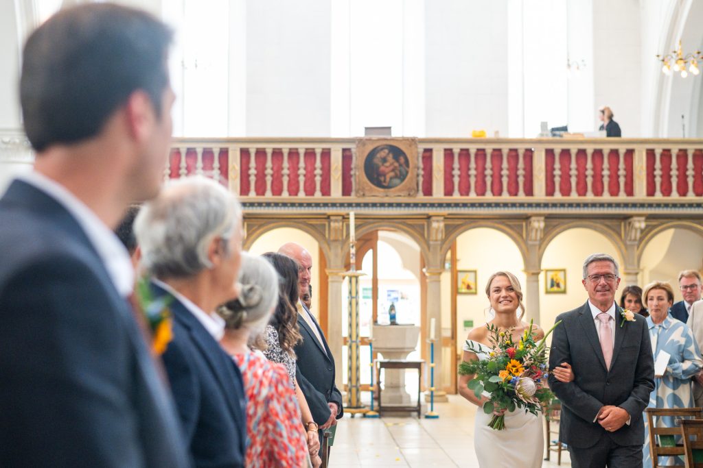 The bride walking down the aisle with her father at St John's Church, Dulwich, smiling towards the groom
