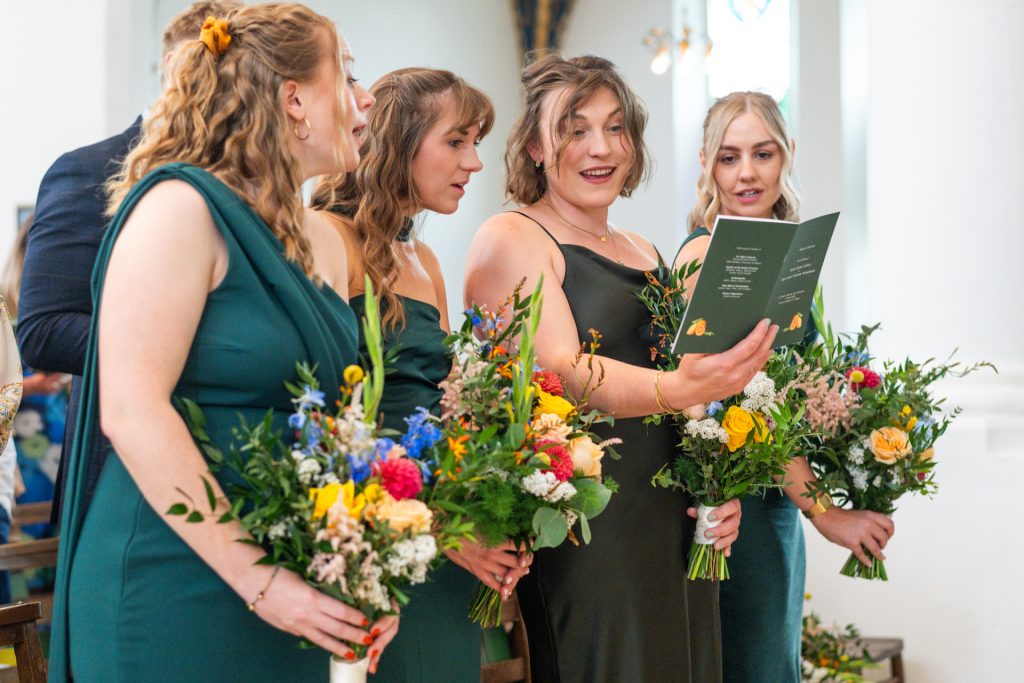 Bridesmaids in green dresses reading the order of service together before the ceremony at St John's Church, Dulwich