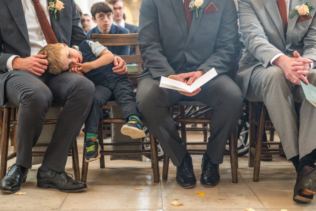 A young boy resting his head on a groomsman's lap during the ceremony at St John's Church, Dulwich
