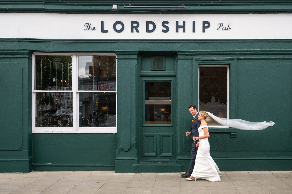 Bride and groom walking past The Lordship Pub's green frontage in Dulwich with champagne, veil catching in the wind