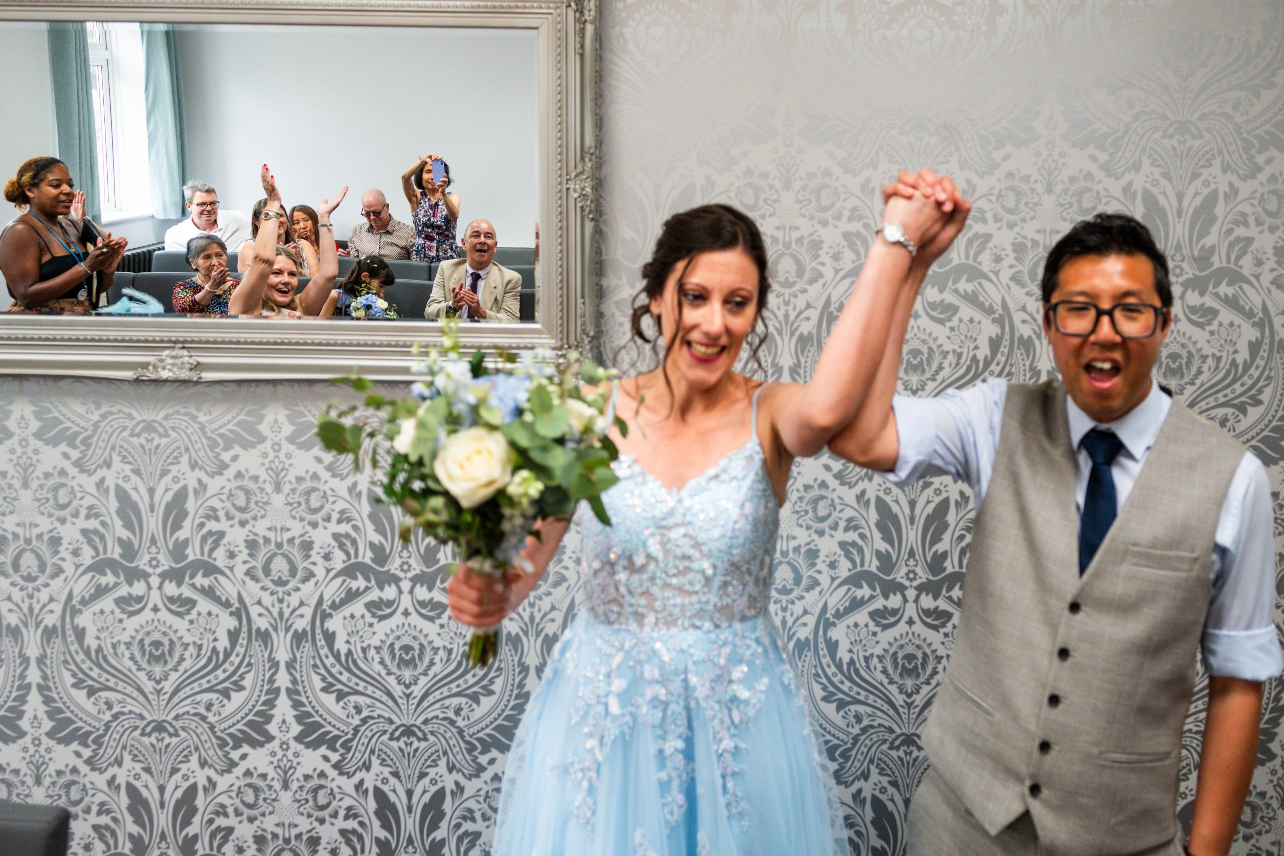 Bride in a pale blue dress and groom raising their joined hands in celebration while guests applaud, reflected in a mirror, London Wedding photographer, documentary wedding photography